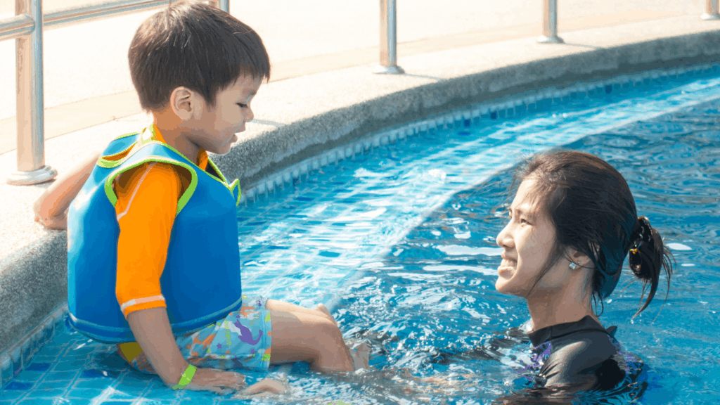 A young boy wearing a blue and orange life vest sits on the edge of a swimming pool, smiling down at a woman in the water who is looking up at him encouragingly.