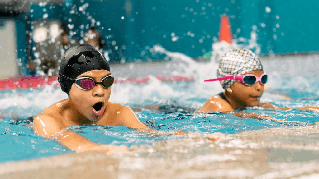 Two children wearing swim caps and goggles swim in a pool during a lesson, with water splashing around them.