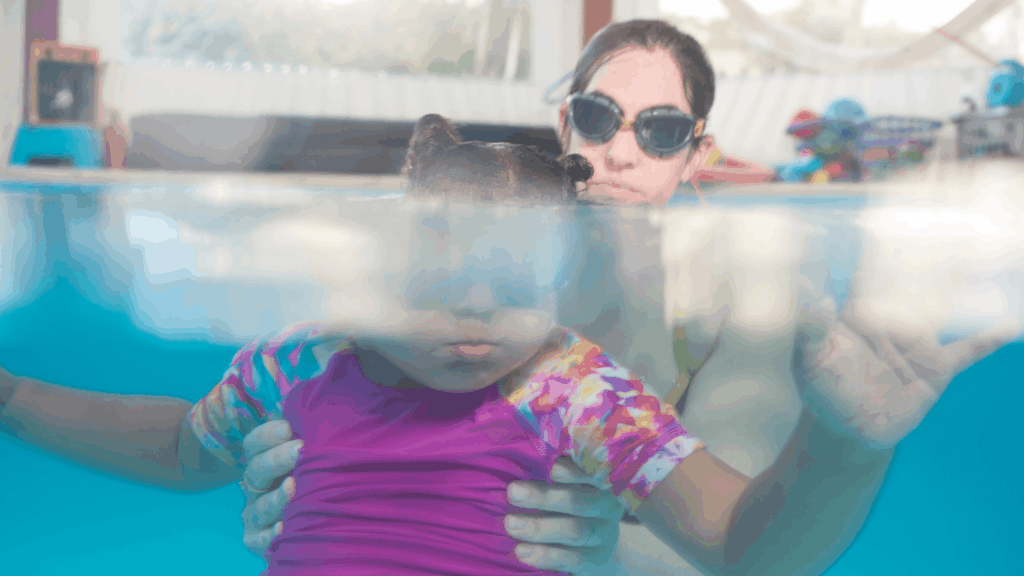 Adult swim instructor supports a young child in a pool during a swimming lesson, viewed partly above and below the waterline.