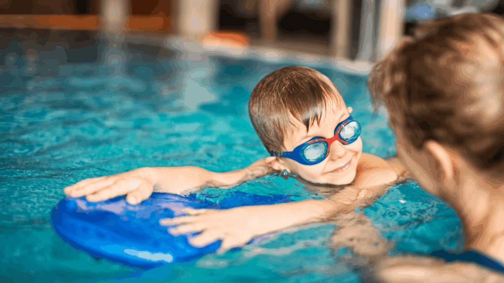 Child wearing swim goggles and holding a kickboard while practicing swimming with an instructor in a pool.