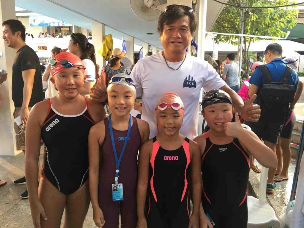 A coach poses with four swimmers in racing suits and caps at a swim meet
