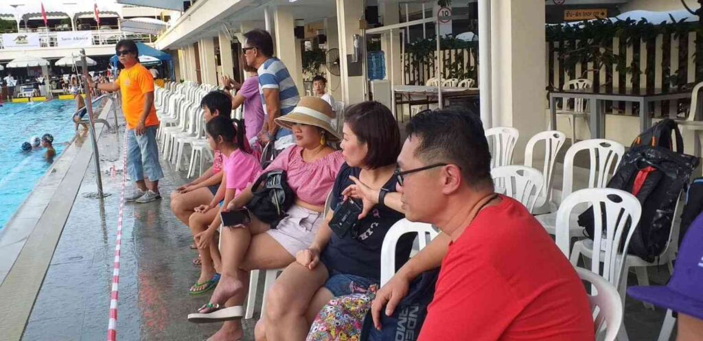 Parents sitting poolside, watching their children swim during a competition.